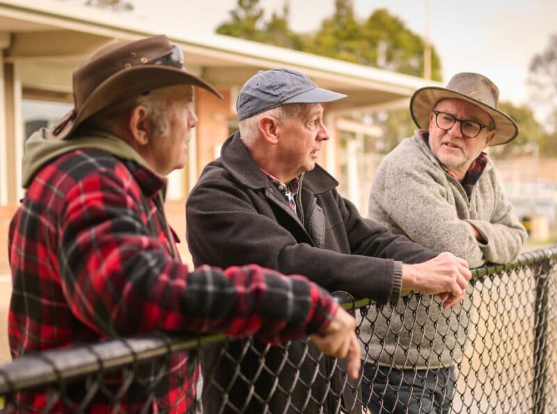 Three men chatting outdoors, leaning against a fence.